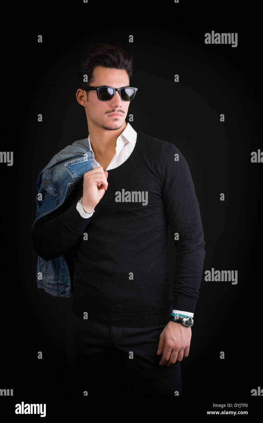 Attraente, alla moda giovane con maglione nero e occhiali da sole su sfondo nero Foto Stock