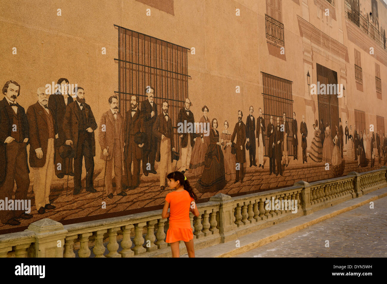 Giovane ragazza cubana in arancione a piedi da un dipinto murale di classe superiore intellettuali dell'epoca coloniale a l'Avana Vecchia Cuba Foto Stock