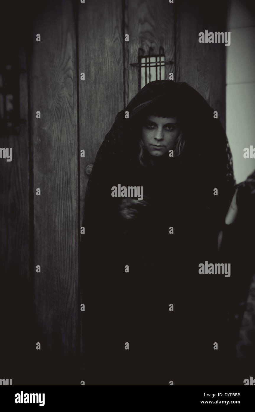 Una strega sorge nelle ombre con una testa di copertura nella parte anteriore di un'annata porta. Foto Stock