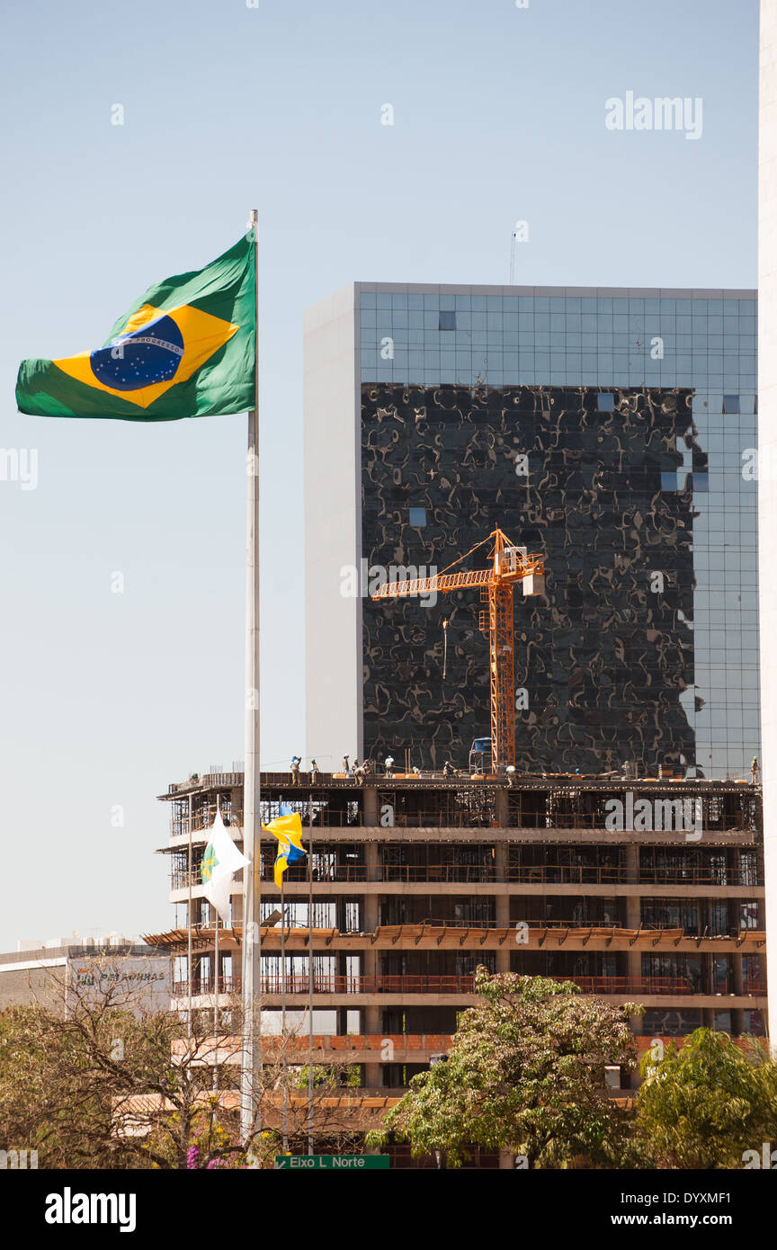 Brasilia, Brasile. Il nuovo edificio in costruzione con la bandiera brasiliana e un vecchio edificio completato con il vetro dello specchio anteriore. Lavoratori sui piani superiori e una gru da cantiere. Foto Stock