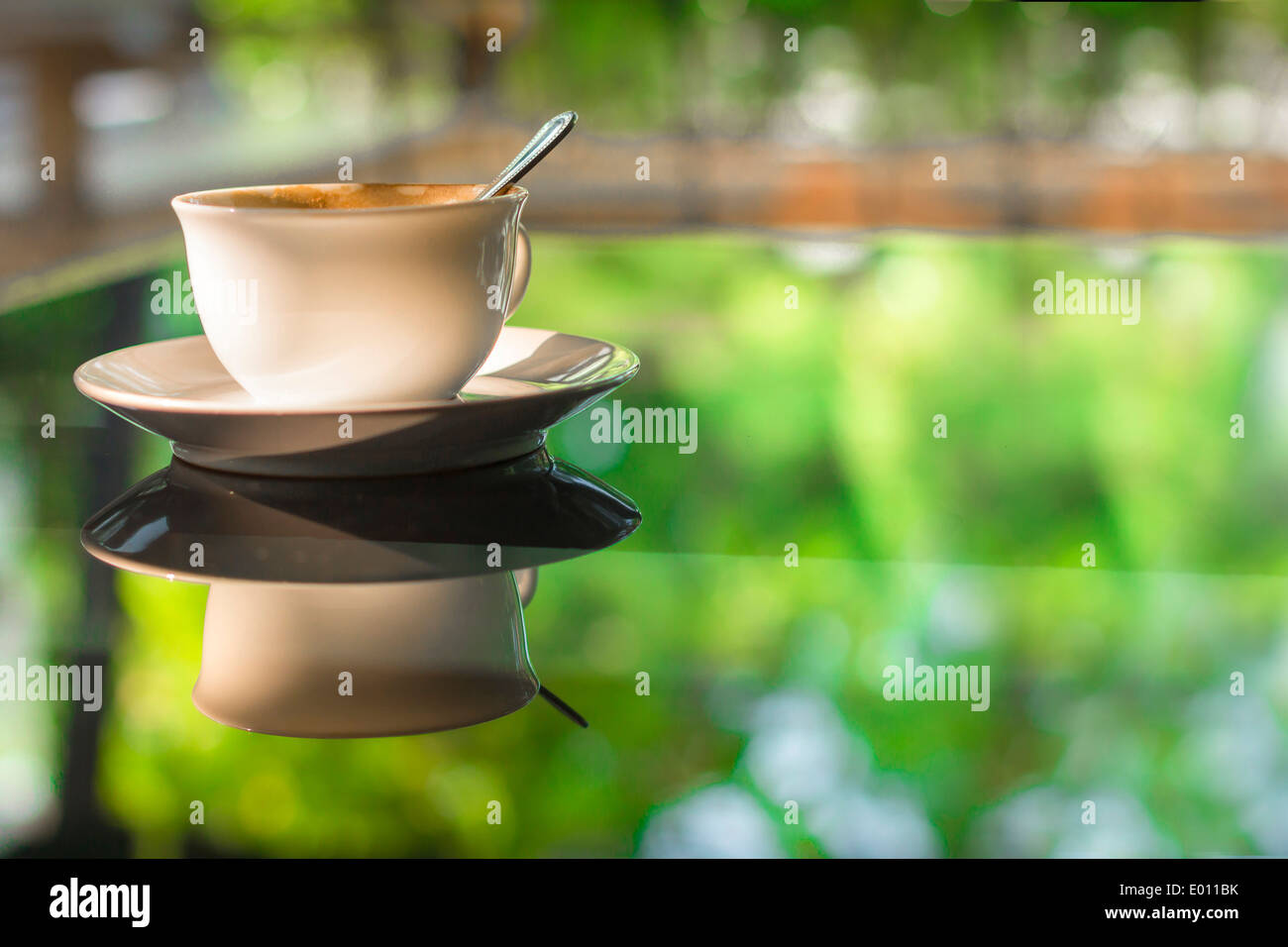 Tazza di caffè sul vetro dello specchio nella tabella riflettono il verde giardino estivo luce del sole di mattina Foto Stock