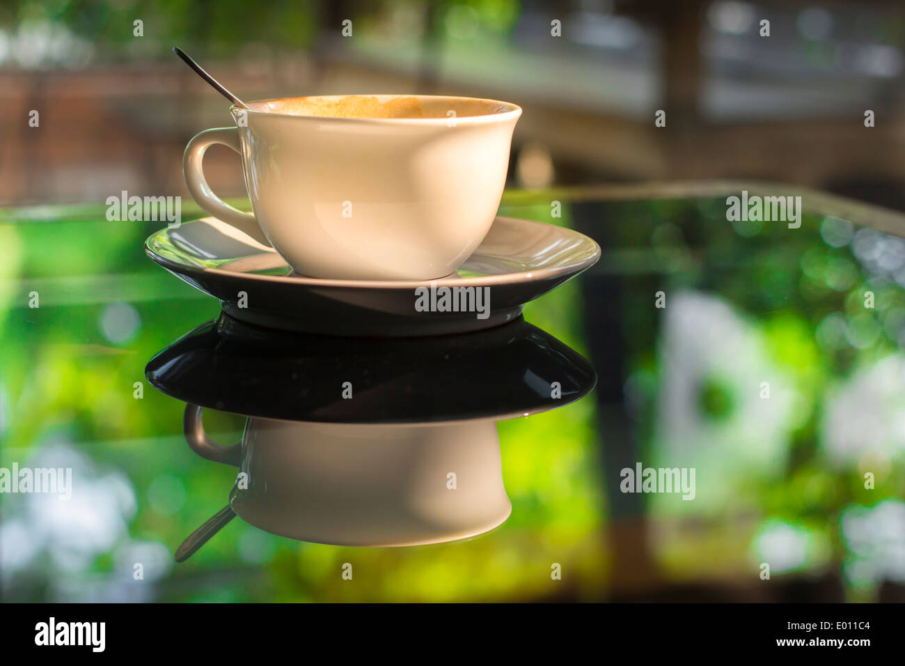 Tazza di caffè sul vetro dello specchio nella tabella riflettono il verde giardino estivo luce del sole di mattina Foto Stock