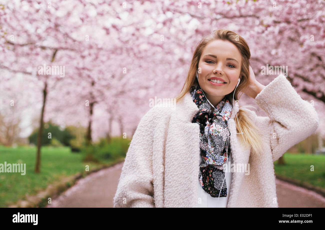Sorridente giovane femmina modello di cuffie da indossare mentre al parco. Bella donna che indossa rivestire a primavera sbocciano i fiori parco. La donna caucasica. Foto Stock