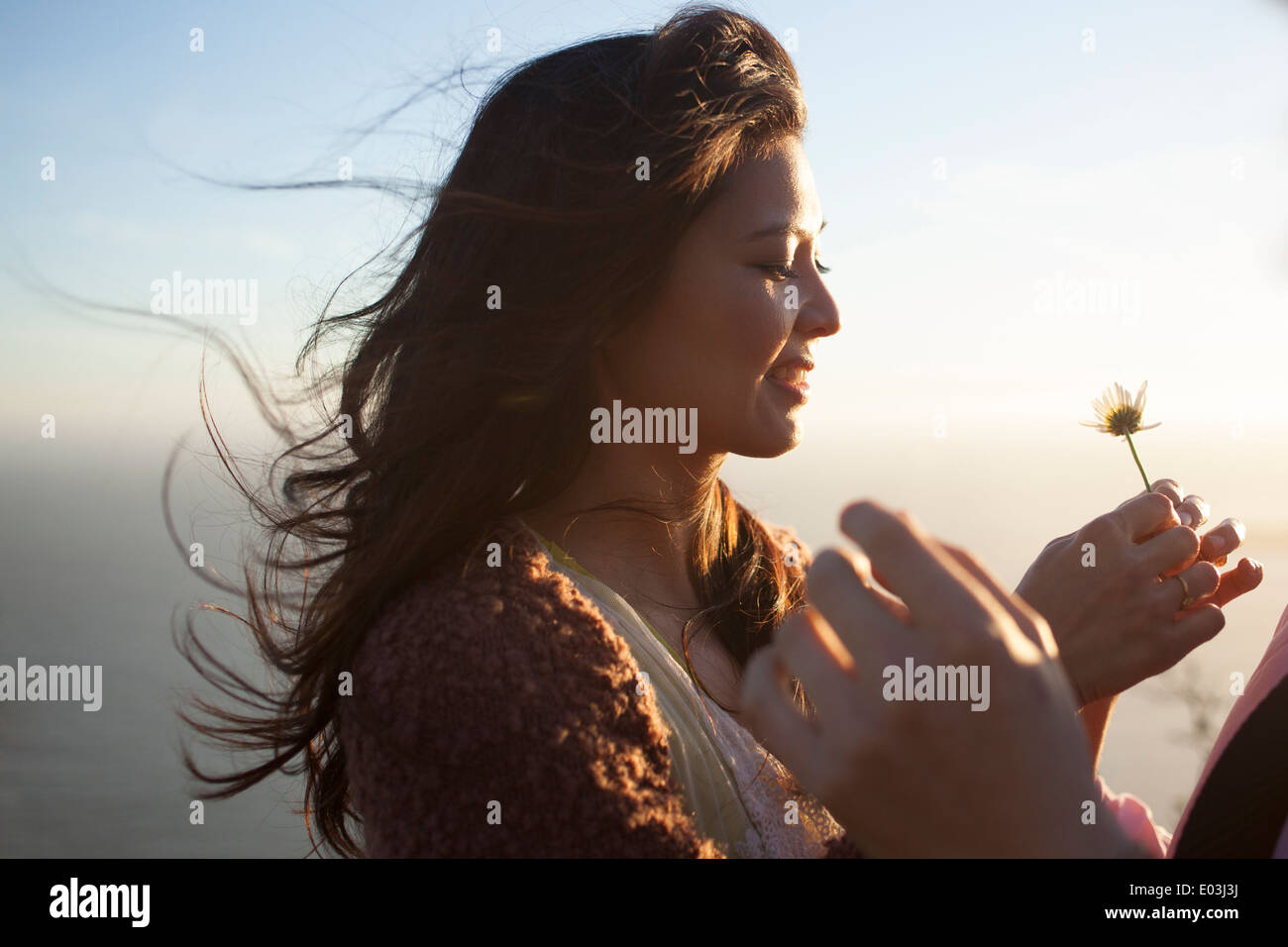 Donna asiatica con battente capelli da una brezza marina e il tramonto dietro di lei con una margherita in mano Foto Stock