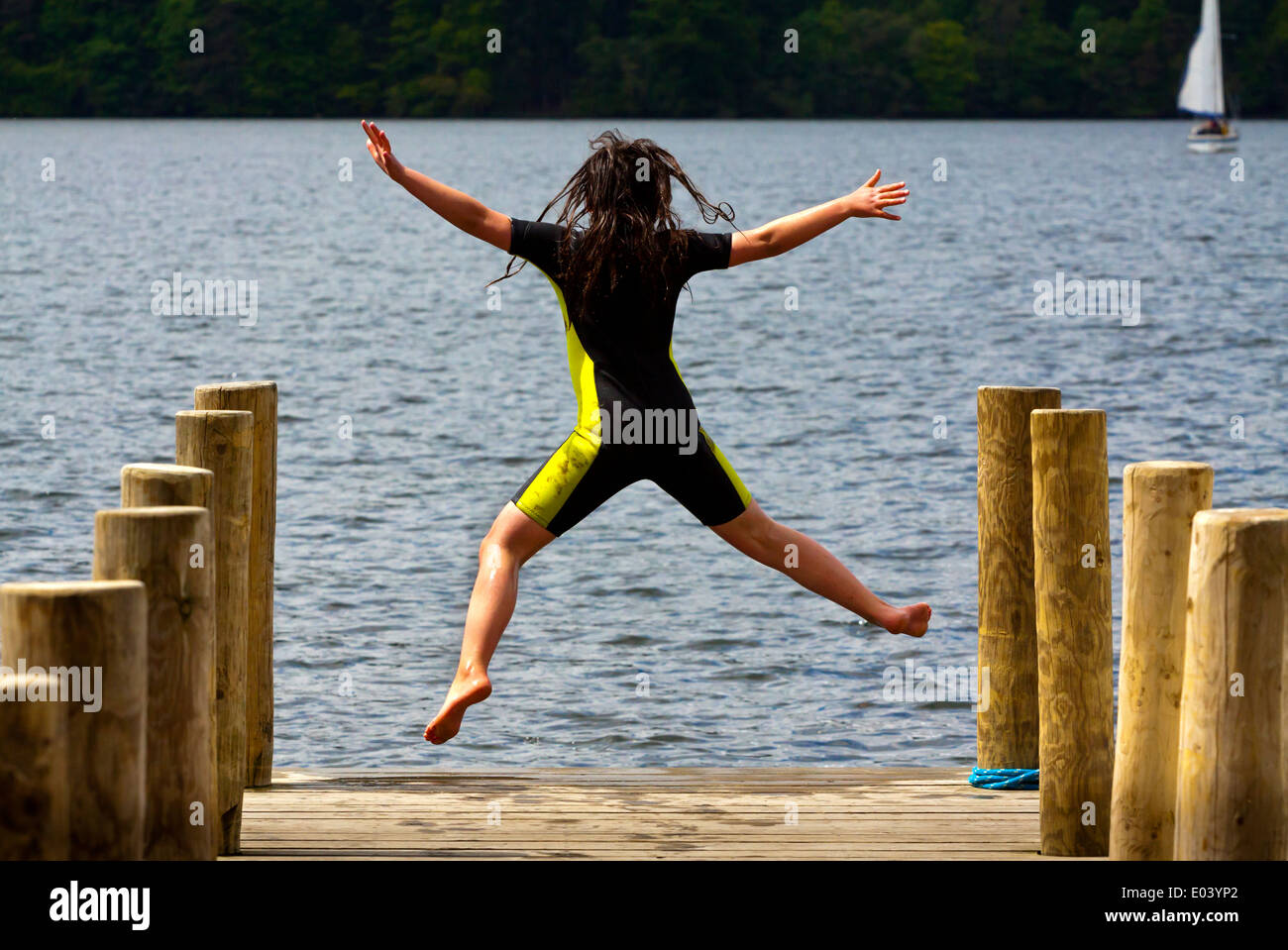 Ragazza di dodici anni in wetsuit jumping dal pontile in legno nel lago di Windermere nel Parco Nazionale del Distretto dei Laghi Cumbria Inghilterra England Regno Unito Foto Stock