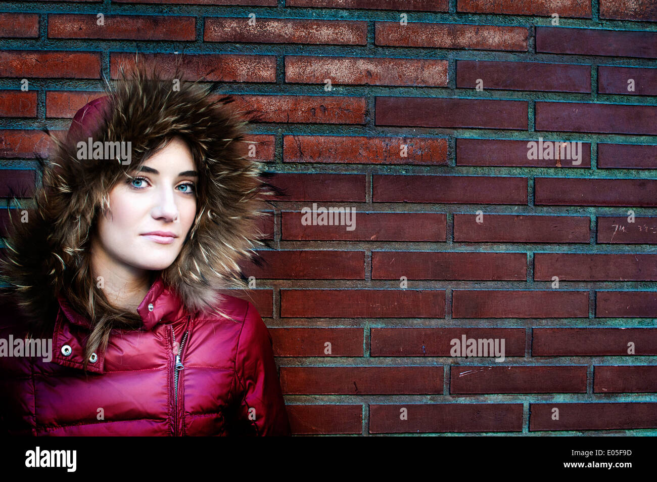 Donna spagnola con gli occhi blu poste contro un muro di mattoni che indossa una rosa con cappuccio cappotto invernale Foto Stock