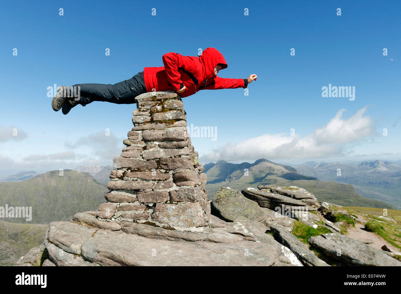 Un hillwalker in una giacca rossa fingendo di essere superman, volare sul vertice cairn di Beinn Alligin in Torridon, Scozia Foto Stock