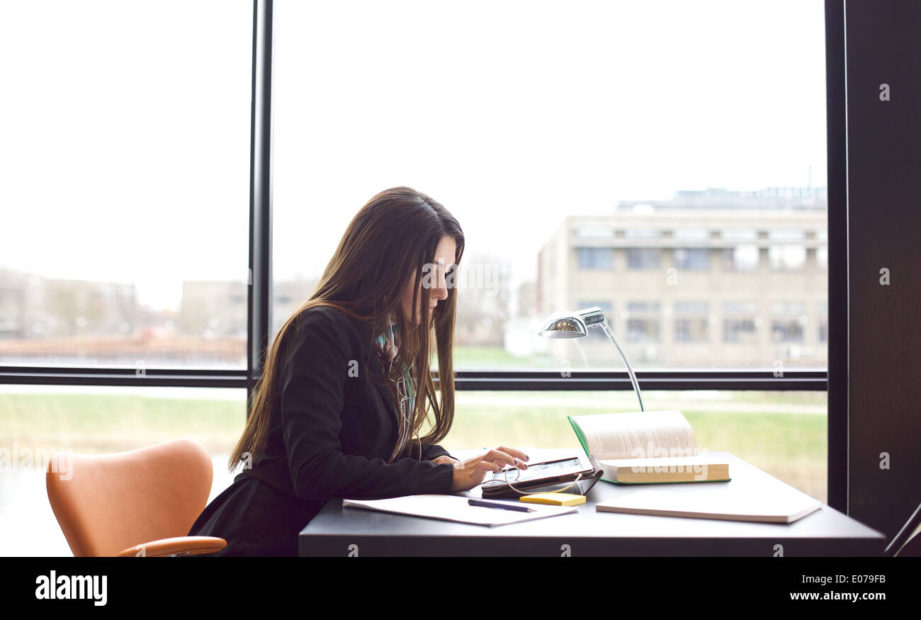 Giovane donna caucasica prendendo appunti dalla tavoletta digitale per il suo studio. Studentessa seduta a tavola con libri e tablet. Foto Stock