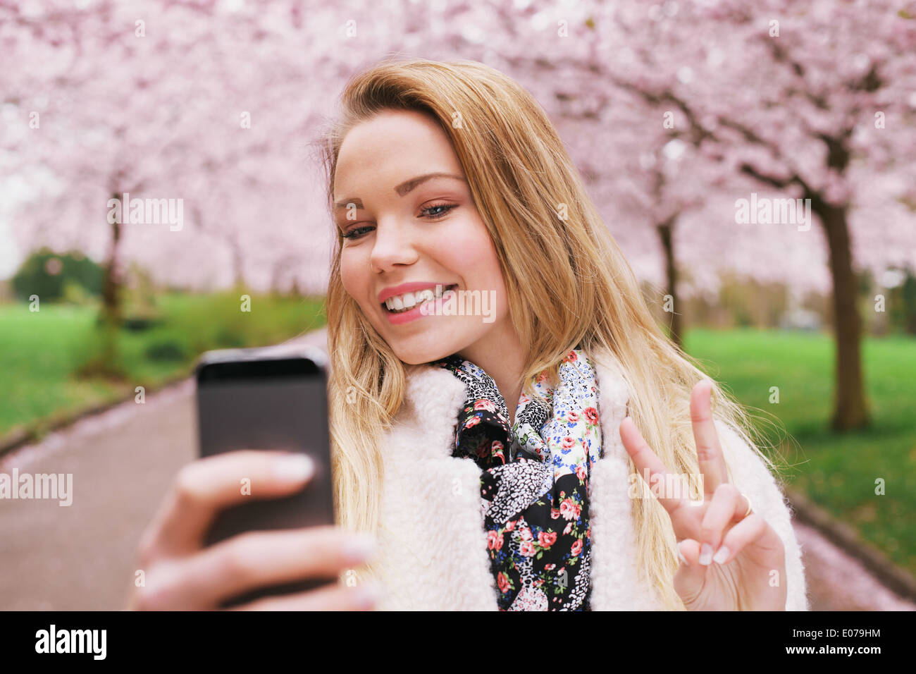 Piuttosto giovane modello femminile gestualità segno di pace mentre prendendo la sua immagine con un telefono cellulare. Bella caucasian giovane donna. Foto Stock