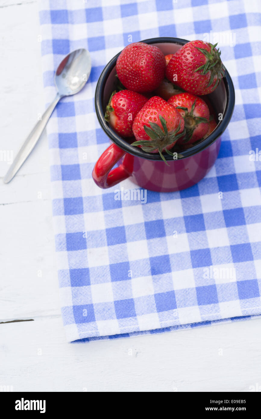 Un piccolo rosso vaso riempito con le fragole e un cucchiaio su un blu e bianco a scacchi in canovaccio. Foto Stock