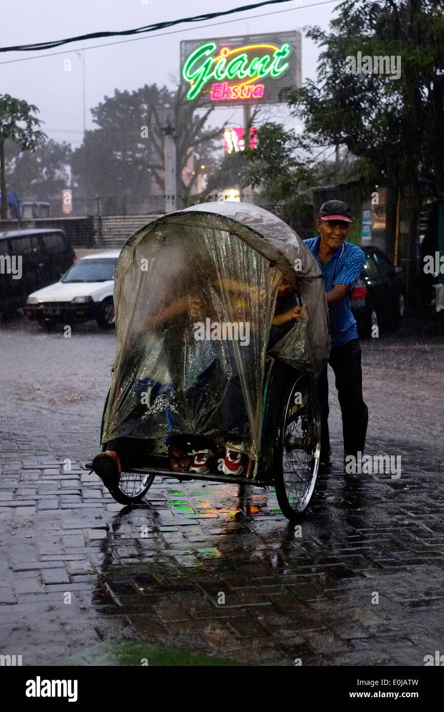 Becak driver e i suoi passeggeri come un diluvio di piogge monsoniche scurisce il tardo pomeriggio sky a malang indonesia Foto Stock