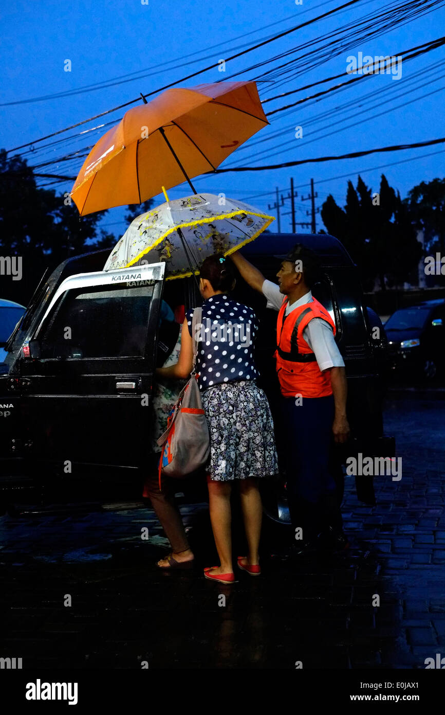 Parcheggio attendant ripara i passeggeri delle auto con un ombrello come un diluvio di piogge monsoniche scurisce il tardo pomeriggio sky a malang Foto Stock