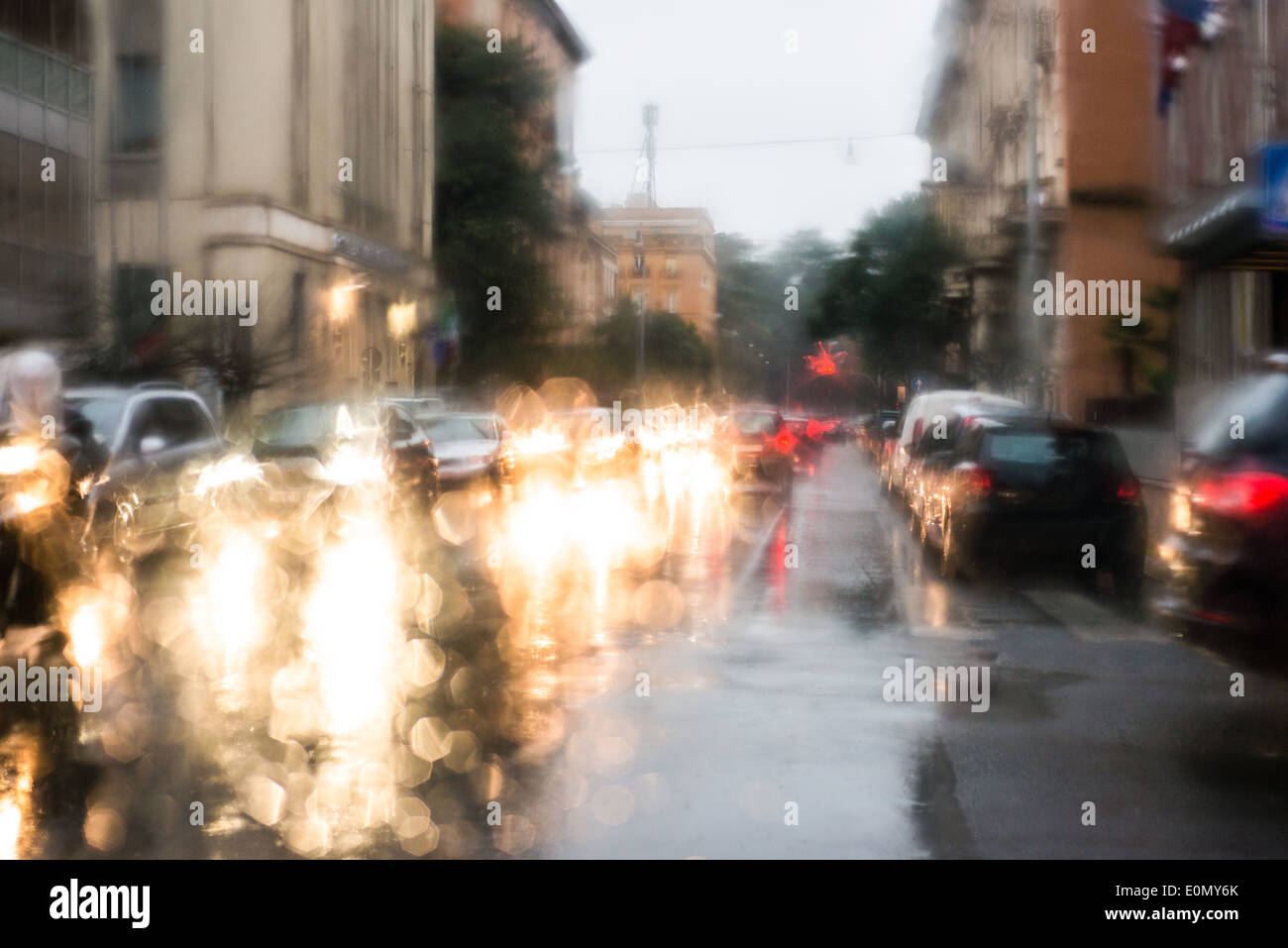 Luce sfocata di vetture visto attraverso un parabrezza bagnato con alcune gocce di pioggia su di esso nella mattina in anticipo di un giorno di pioggia Foto Stock