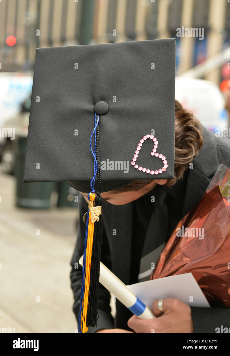 Manhattan, New York, Stati Uniti - 21 Maggio 2014 - sulla settima avenue, un giovane uomo che si è laureato dall'università di passo che giorno si inclina la sua testa per mostrare la parte superiore della sua graduazione nera scheda di mortaio hat, che è decorato con strass rosa cuore di Manhattan. Un blu e oro infiorescenza staminifera topper è consegnato dal cappello. Foto Stock