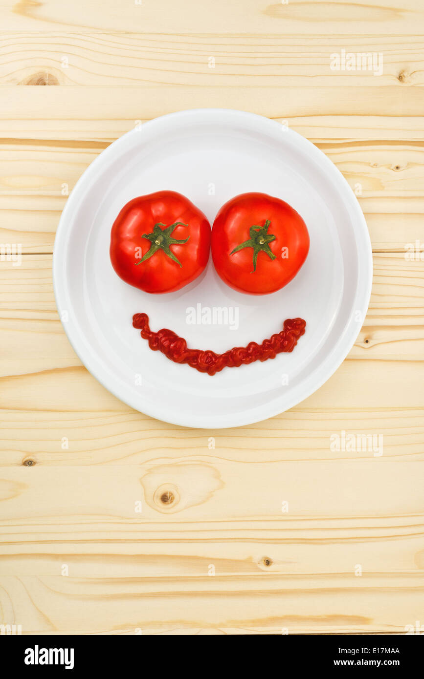 Divertimento e alimenti per bambini - pomodori e ketchup rendendo Smiley face servita su una piastra bianca sul tavolo di legno. Foto Stock