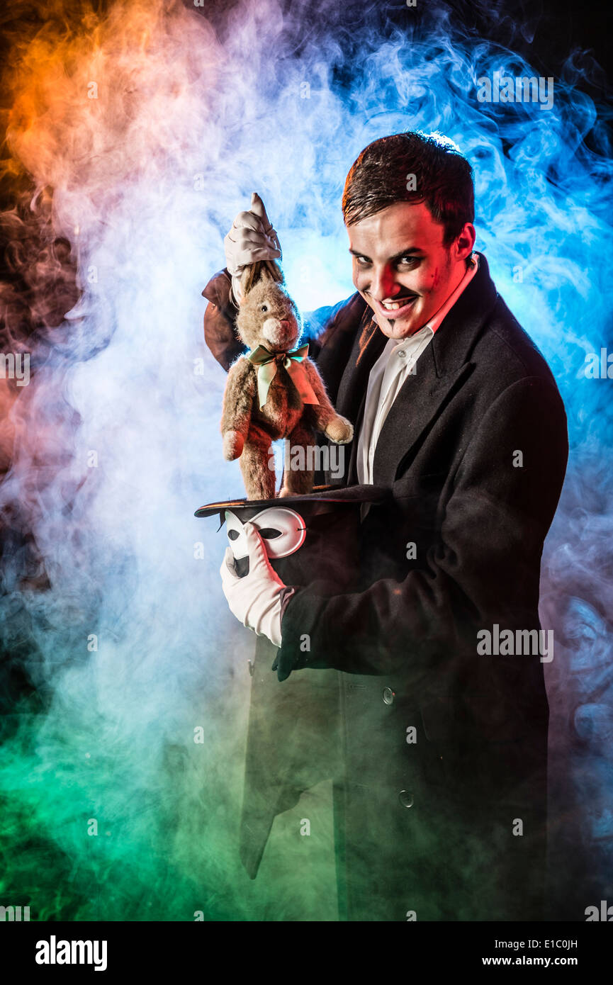 Un male sinister mago cattivo inghirlandato in fumo tirando un bambino giocattolo morbido il coniglio fuori da un cappello a cilindro Foto Stock