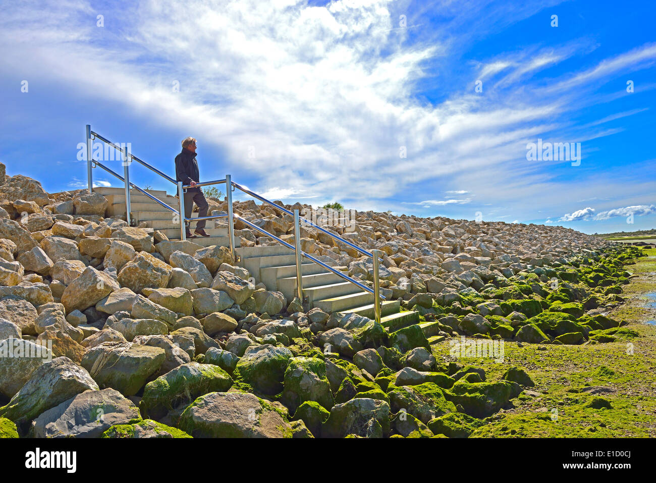 Uomo a piedi giù i gradini sulle difese marine della banca di pietra di recente costruzione con formatio di nuvola insolita a West Wittering, vicino a Chichester, West Sussex, Inghilterra, Regno Unito Foto Stock