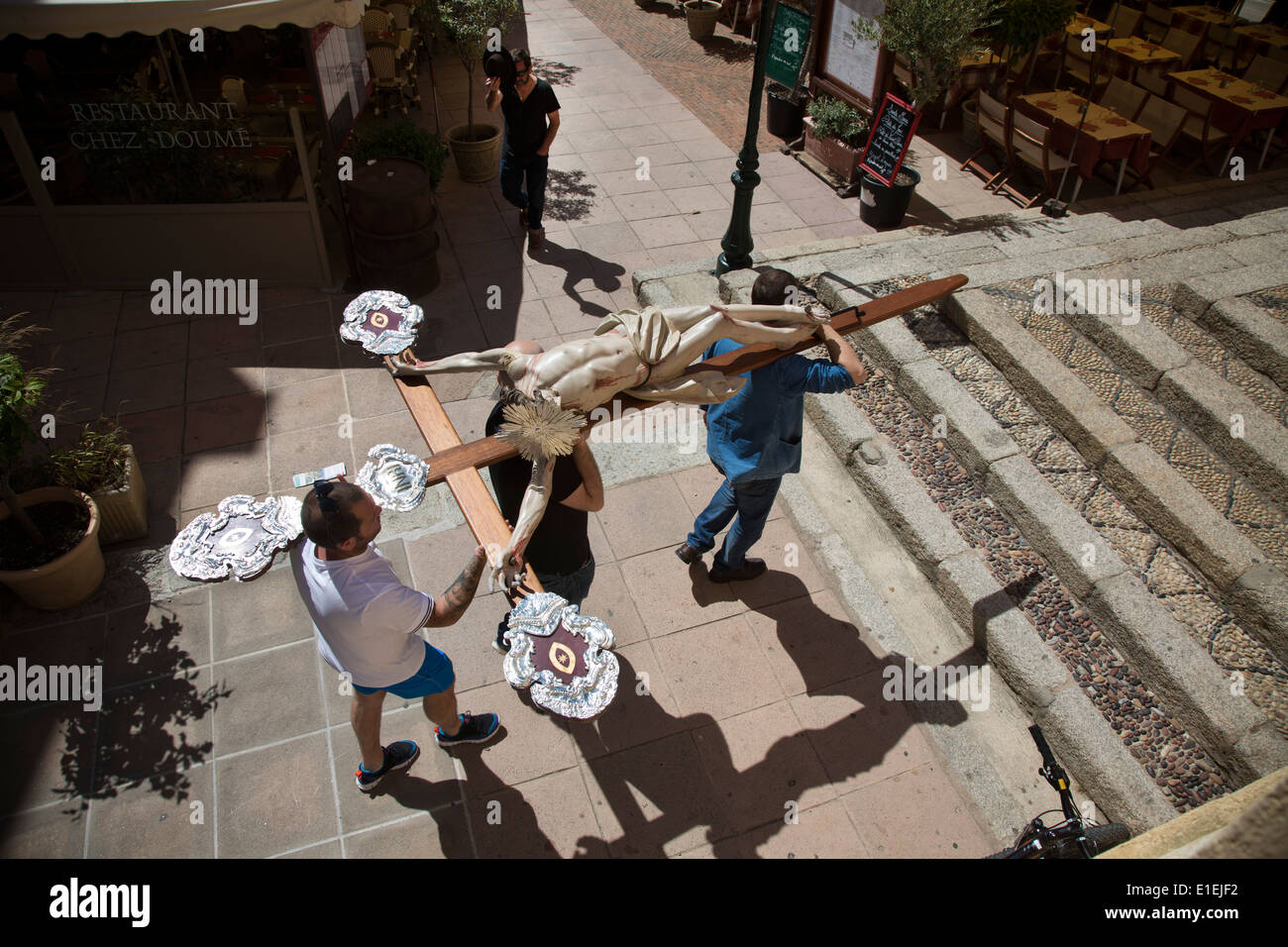 Un uomo suggerimenti il suo cappello a tre uomini con la statua del Cristo sulla croce attraverso le strade della città di Calvi, in Corsica. Foto Stock