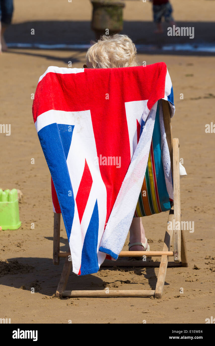 Donna seduta sulla sedia a sdraio sulla spiaggia con Unione Jack asciugamano poggiata sulla sedia a sdraio Foto Stock