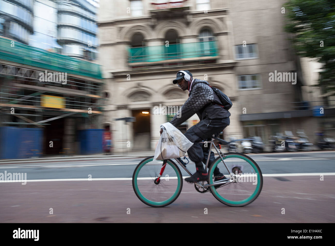 Uomo che indossa le cuffie sopra il suo cappello da baseball, a cavallo di una bicicletta con un blu brillante ruote lungo una strada di Londra. Foto Stock