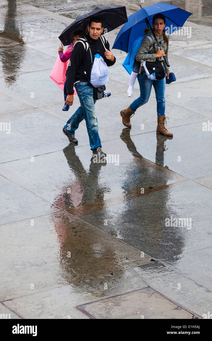Un paio portano ciascuno un bambino ogni sulle loro spalle mentre passeggiate attraverso un wet Trafalgar Square mentre il ricovero sotto gli ombrelloni. Foto Stock