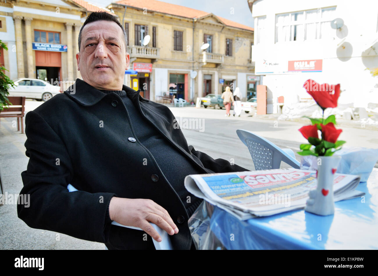 Uomo si siede presso il cafe' tabella sulla strada, leggendo il giornale in Lefka, la parte settentrionale di Cipro Foto Stock