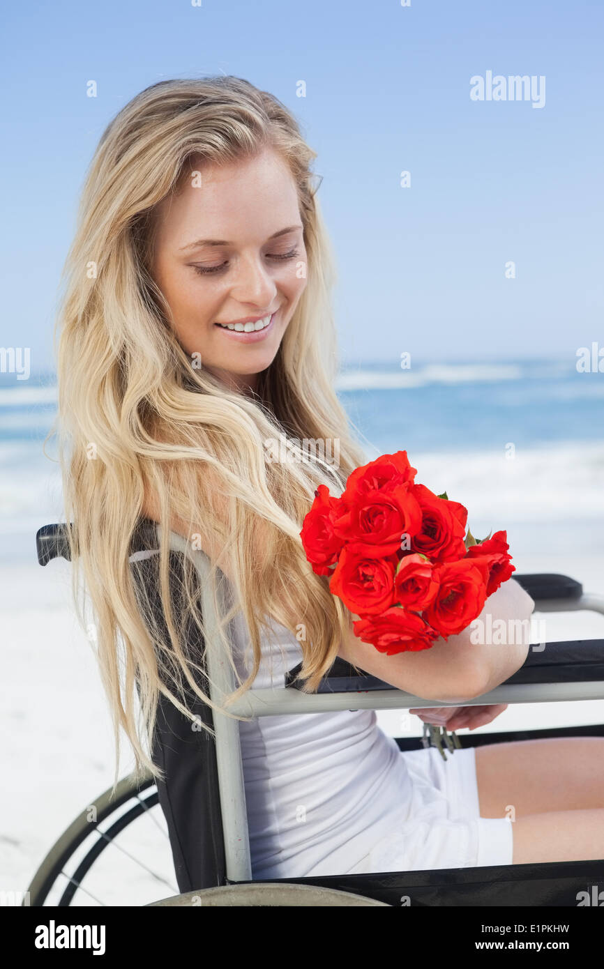 Sedia a rotelle legato bionda sorridente sulla spiaggia tenendo le rose Foto Stock