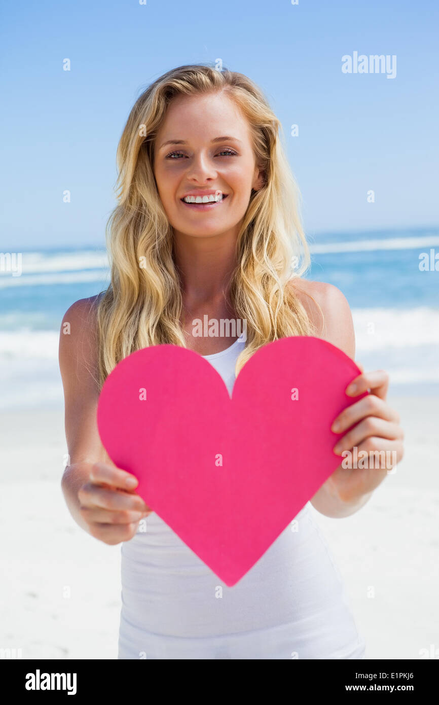 Bionda sorridente mostra cuore rosa sulla spiaggia Foto Stock