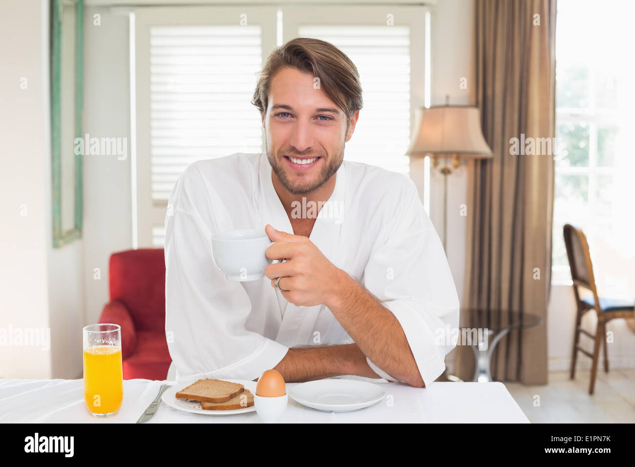 Uomo bello avere la prima colazione nel suo accappatoio sorridente in telecamera Foto Stock