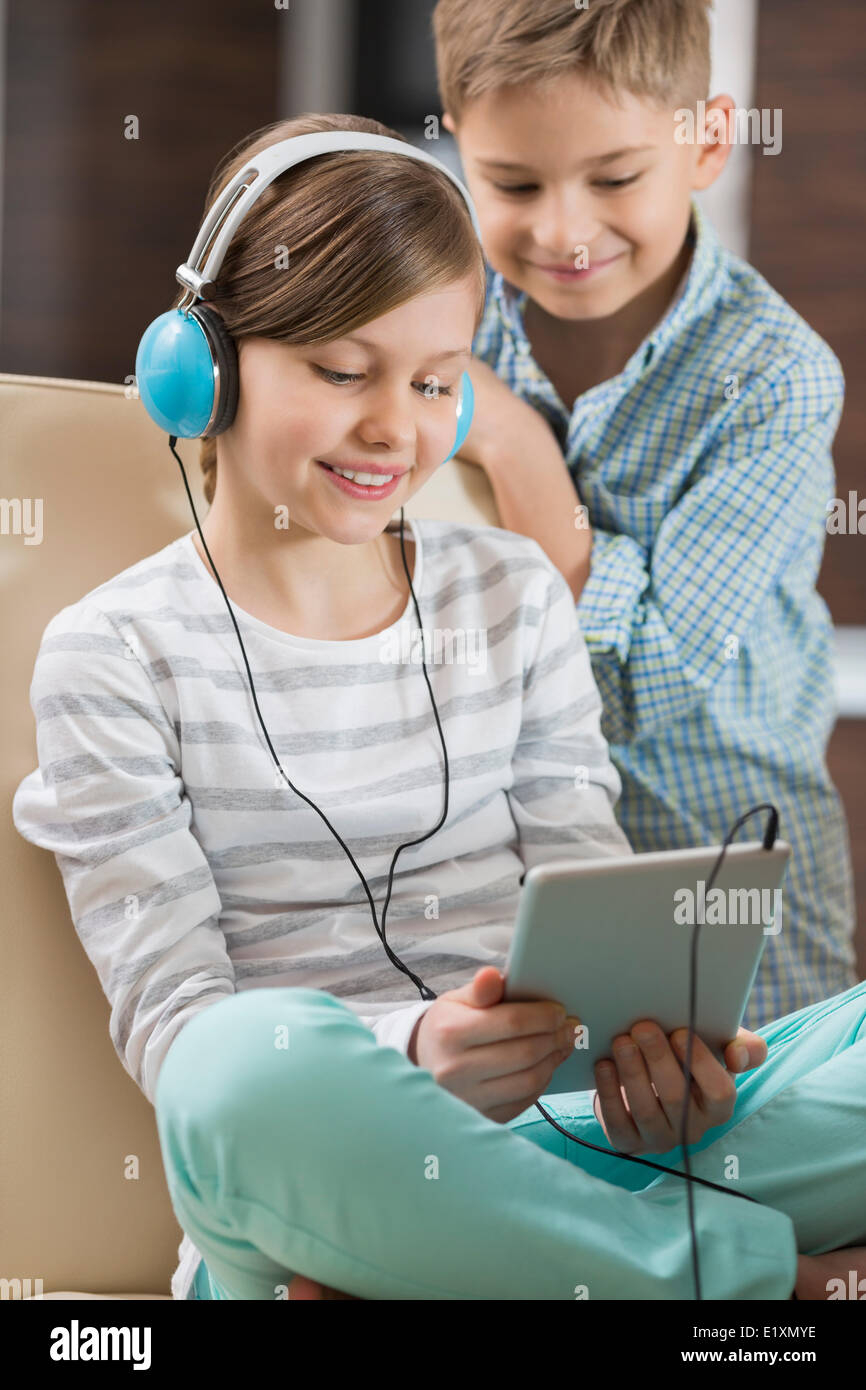 Ragazza carina ascoltando musica su tavoletta digitale mentre il fratello in piedi dietro di lei a casa Foto Stock