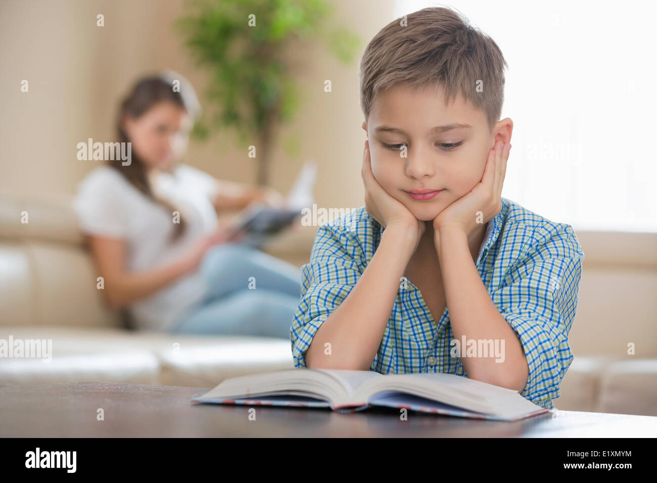 Ragazzo lettura prenota con la madre in background a casa Foto Stock