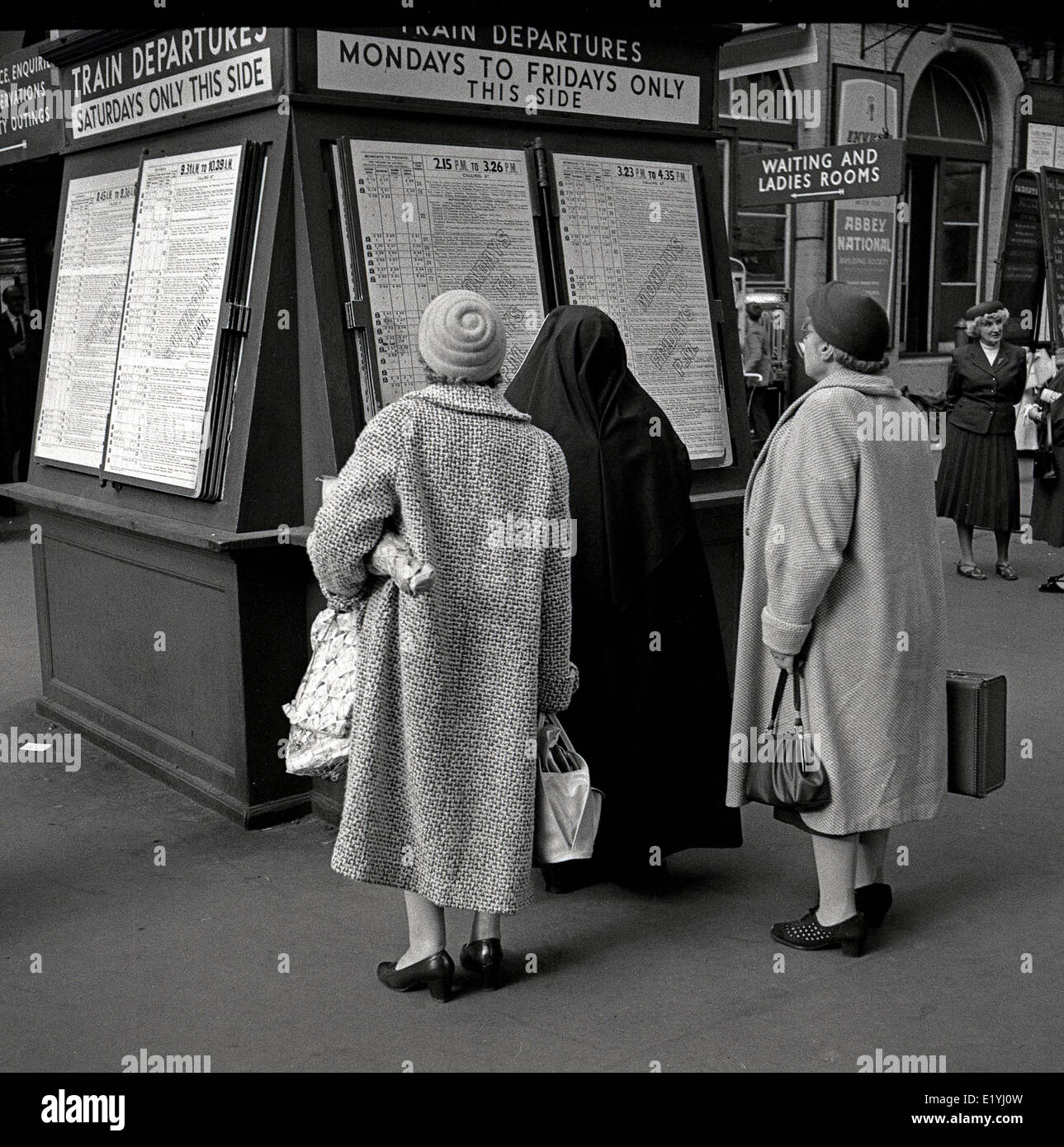 Anni Cinquanta foto storiche, tre donne anziane in cappotti e cappelli, uno con un velo nero, guardando il calendario informazioni su un treno Partenze pensione presso la stazione di Waterloo, Londra. Foto Stock