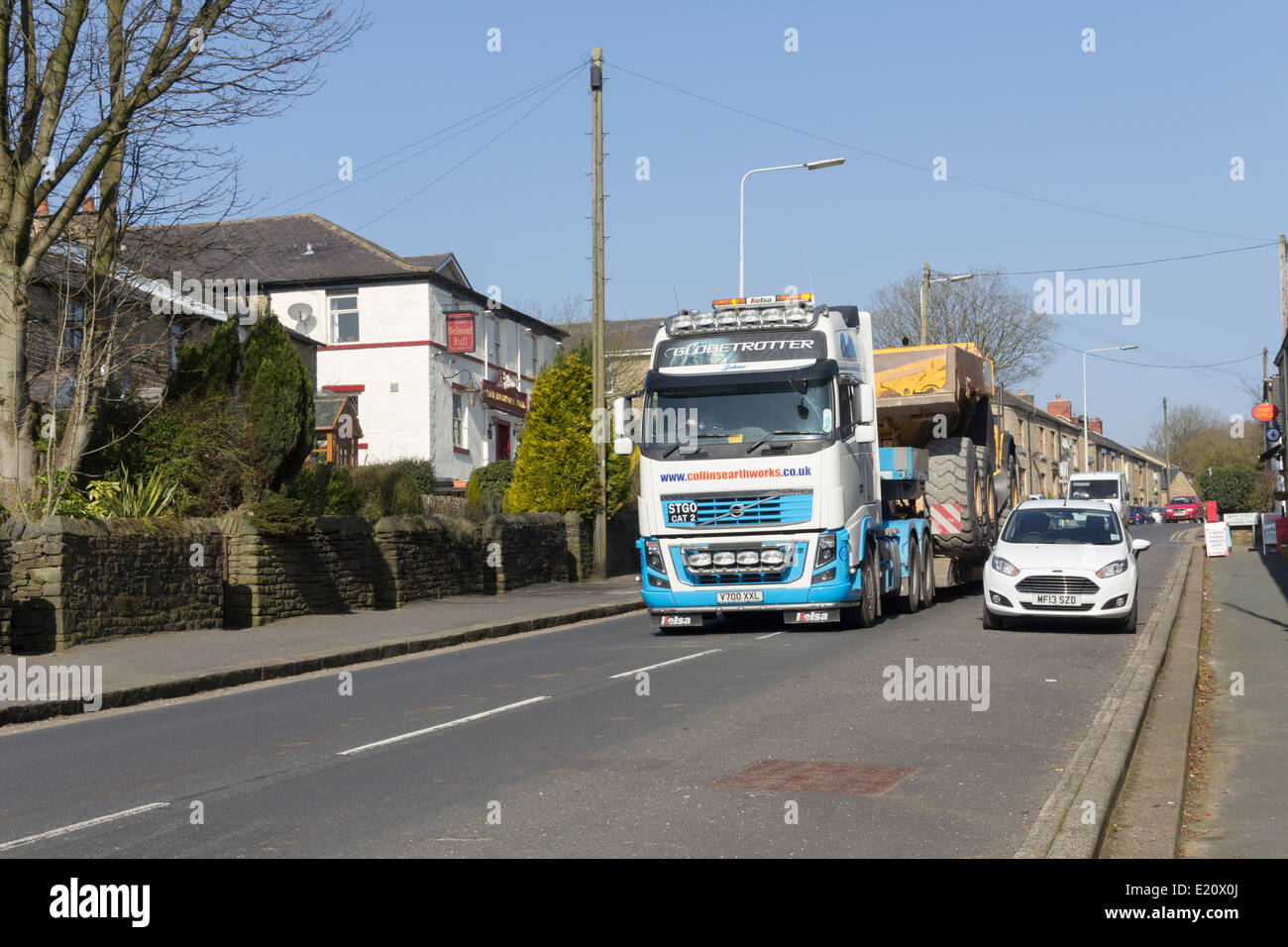 Ampia-carico, movimento terra camion vengono trasportati su una Volvo FH16 bassa-caricatrice attraverso un villaggio nel nord-ovest dell'Inghilterra. Foto Stock