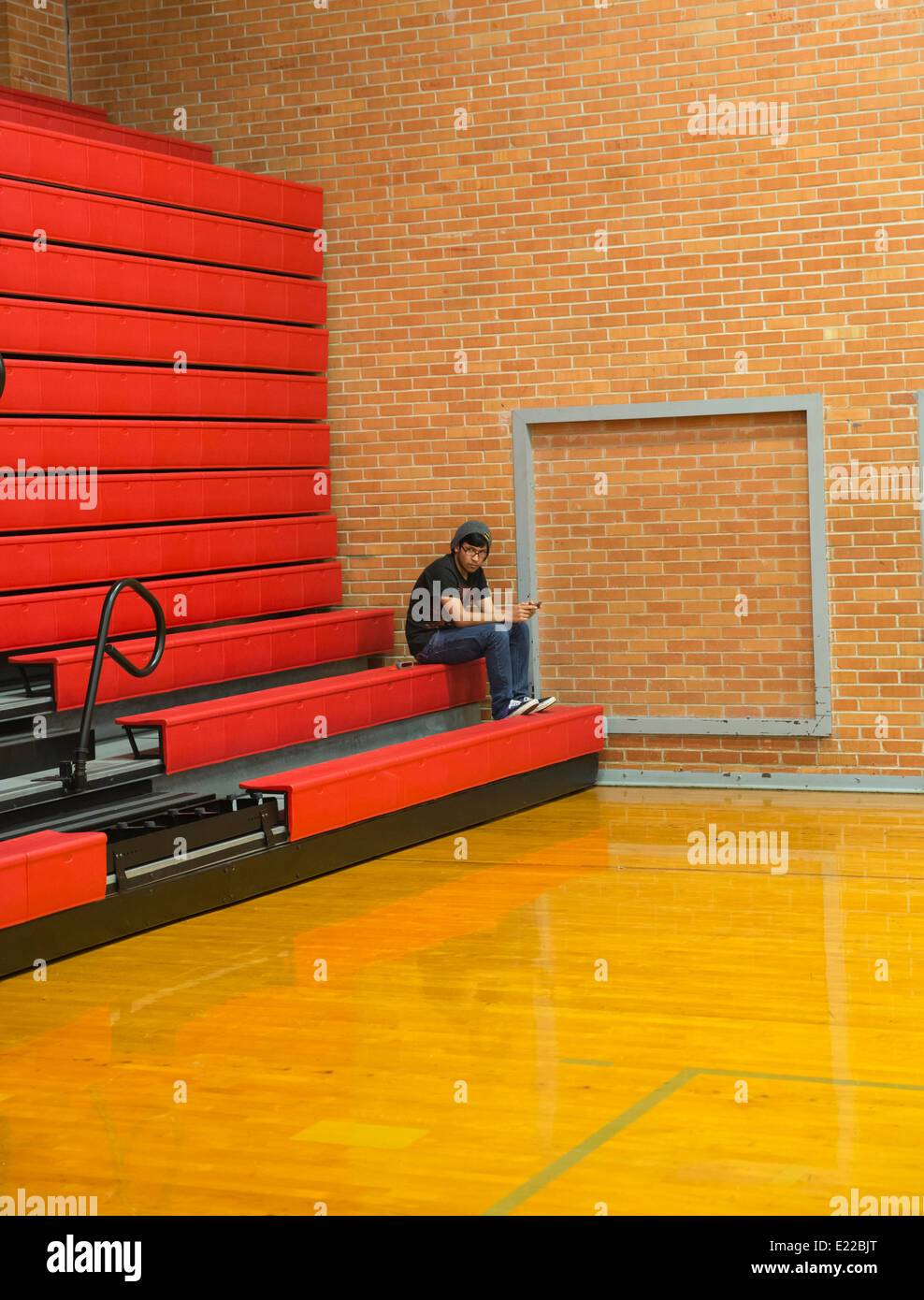Ispanico high school anziano studente utilizza il cellulare per il testo come si siede in tribuna all'interno della palestra della scuola di Austin in Texas Foto Stock