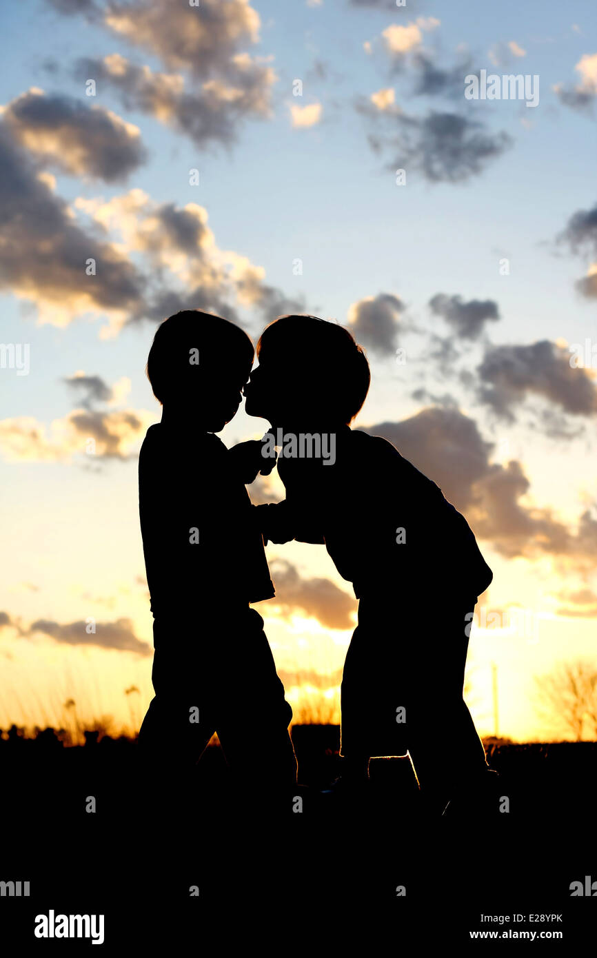 Una silhouette di un giovane bambino baciare il suo fratellino sulla guancia al di fuori al tramonto Foto Stock