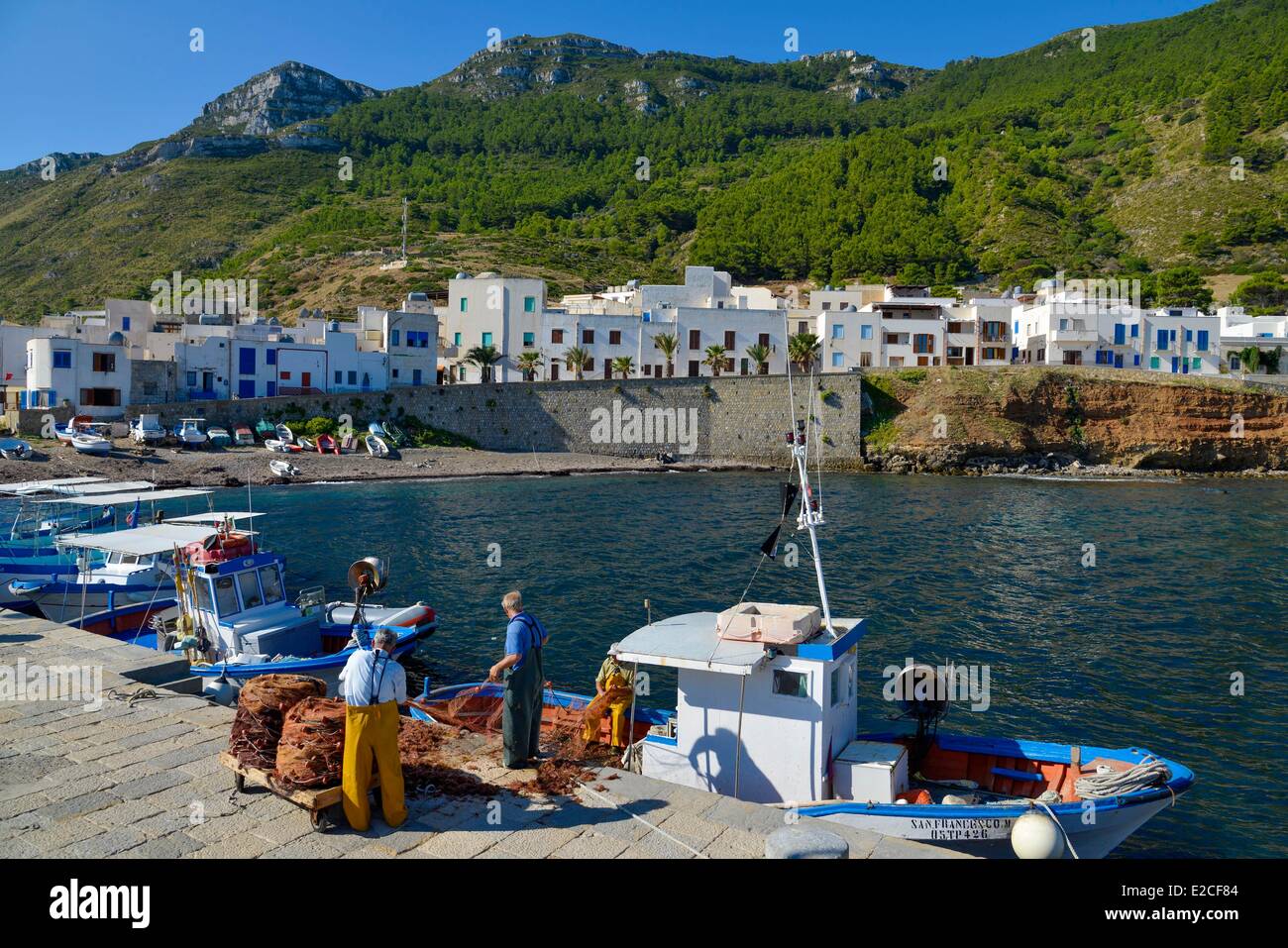 L'Italia, sicilia, isole Egadi, isola di Marettimo, porto di pescatori, reti da pesca unraveling prima della loro partenza per mare con il bianco villaggio con case in background Foto Stock