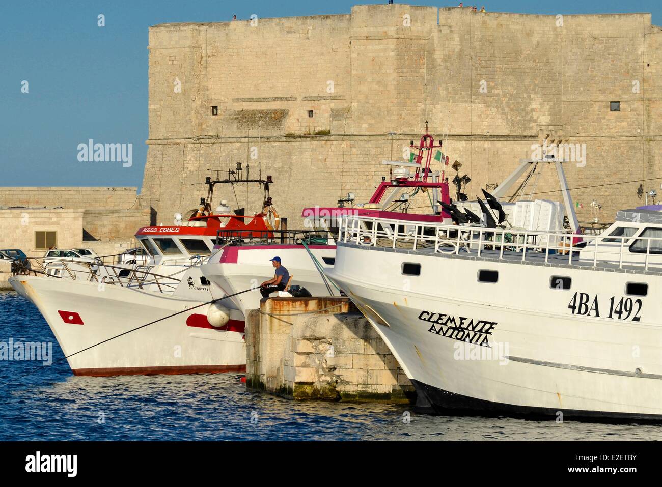Italia Puglia Provincia di Bari Monopoli pescatore seduto su una diga tra due navi per la pesca a strascico con il Fort Santa Maria del XVI secolo in Foto Stock