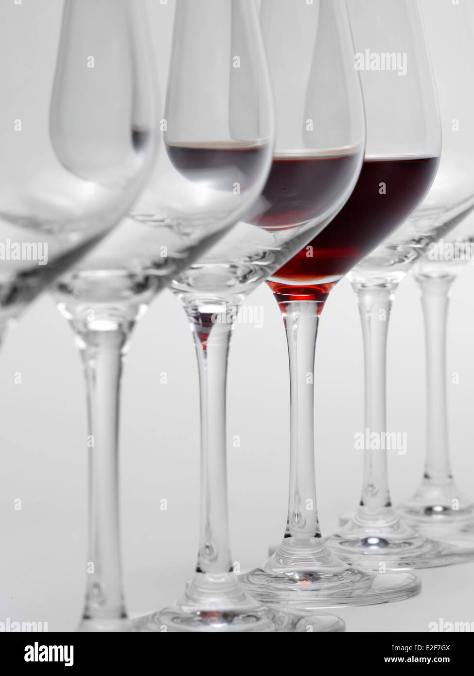 Alcuni bicchieri di vino in piedi consecutivi in una fila in grigio chiaro indietro, uno parzialmente riempito con un bicchiere di vino rosso Foto Stock