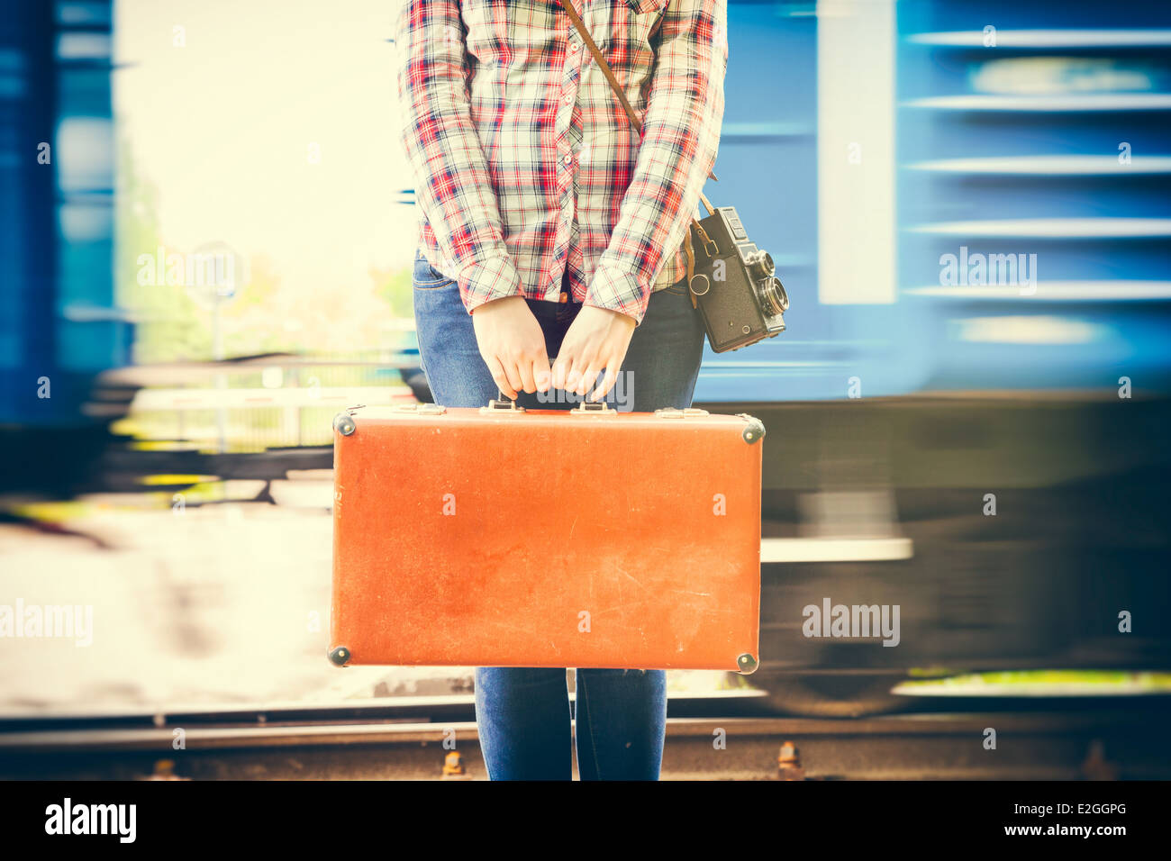 Giovane donna con valigia retrò e fotocamera su piattaforma ferroviaria, vagoni del treno sullo sfondo Foto Stock