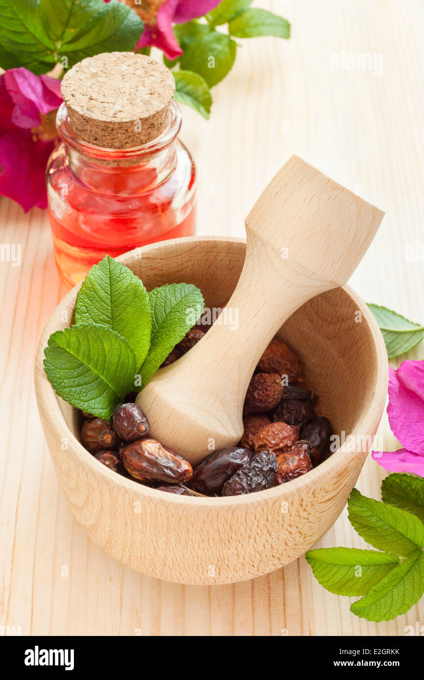 Olio essenziale in una bottiglia di vetro, rosa appassita-hip bacche in mortaio di legno e di rosa mosqueta fiori sul tavolo Foto Stock
