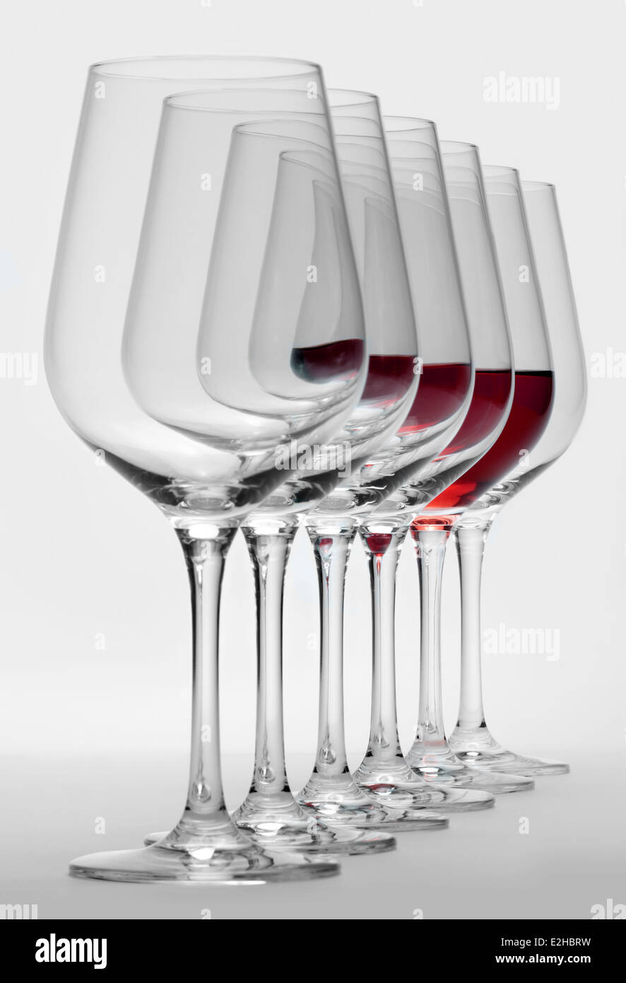 Alcuni bicchieri di vino in piedi consecutivi in una fila in grigio chiaro indietro, uno parzialmente riempito con un bicchiere di vino rosso Foto Stock