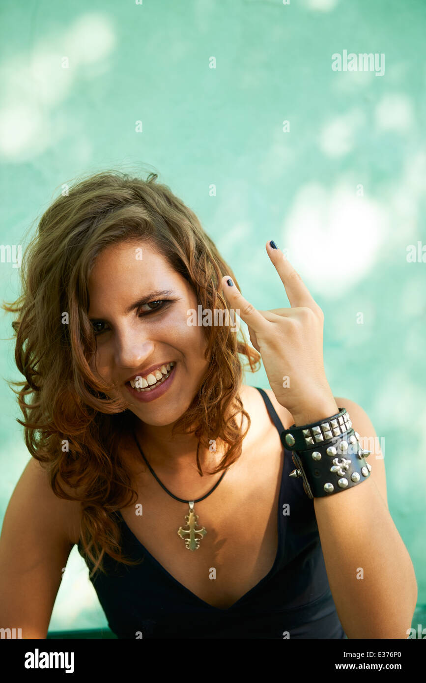 Ritratto di giovane donna rocker gesti con le mani e guardando la telecamera contro il muro verde Foto Stock