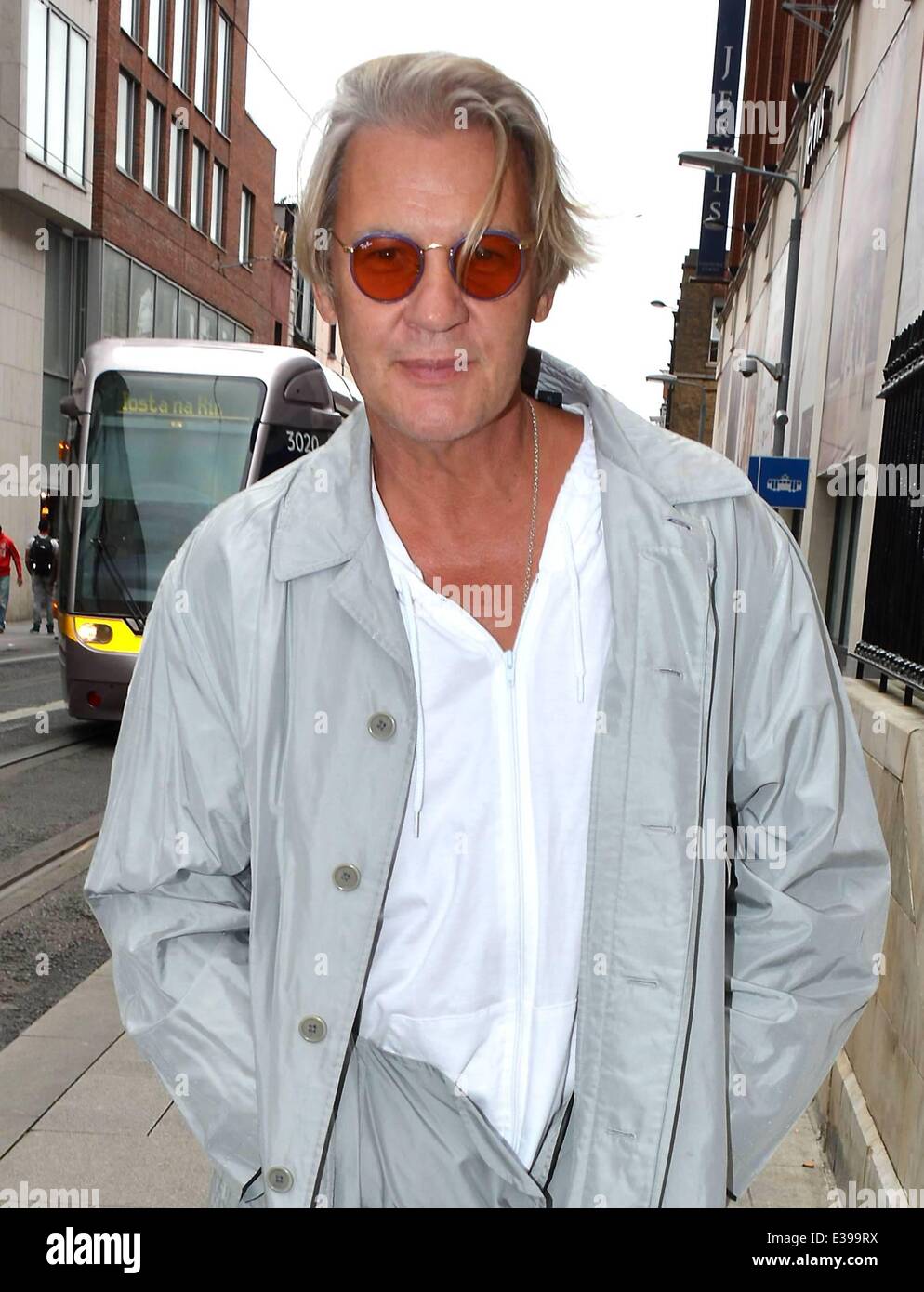 La moda consapevole doppio vincitore Eurovisione Johnny Logan visto oltrepassando il Luas a Jervis Street Shopping Center di indossare occhiali con lenti di colore arancione, grigio di un trench coat, e baby-blu slip-on scarpe senza calze con: Johnny Logan dove: Dublino, Irlanda quando: 27 Ago 2013 Foto Stock