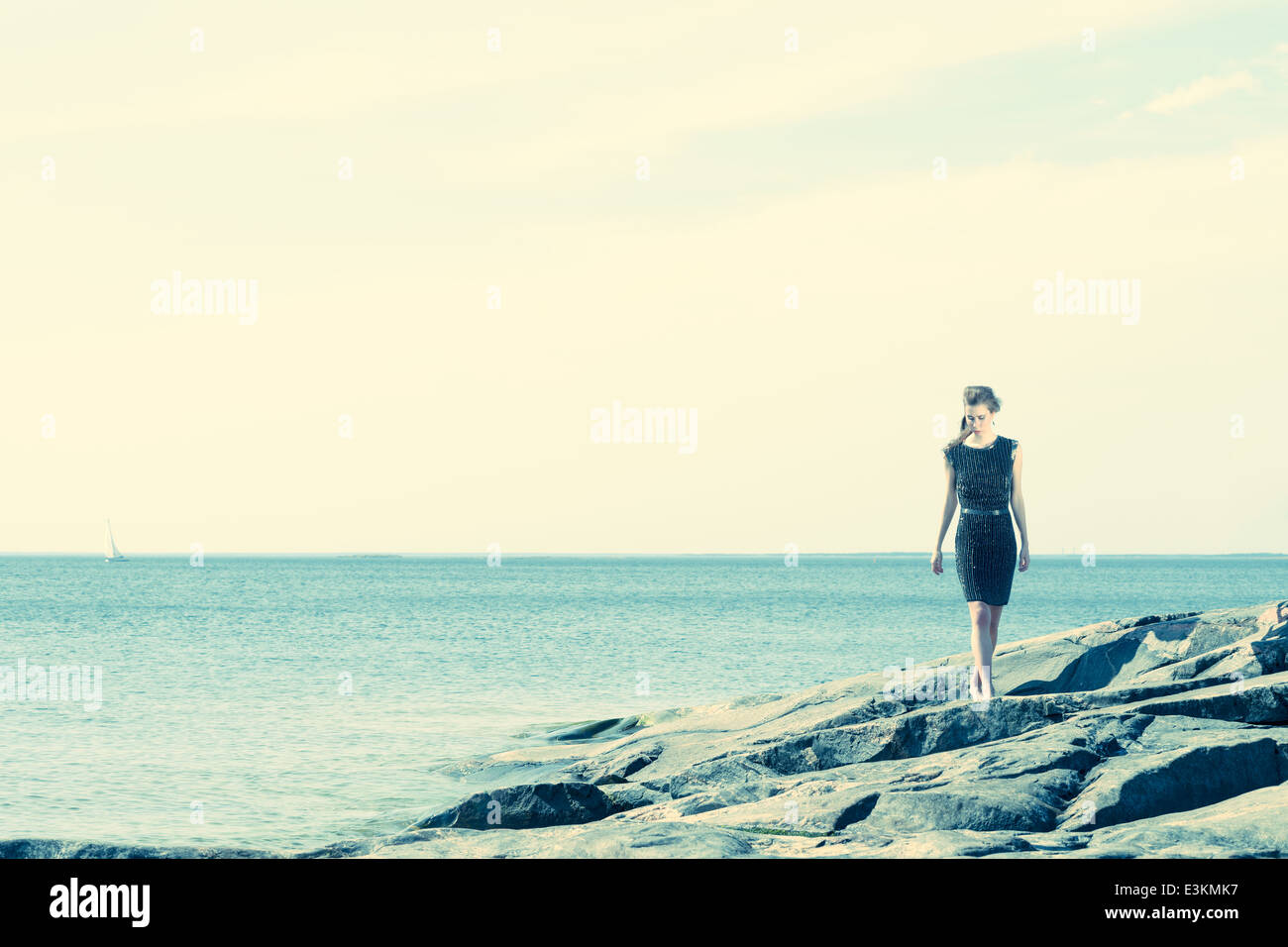 Moda Donna camminando sulla spiaggia rocciosa, la barca a vela e orizzonte sullo sfondo Foto Stock