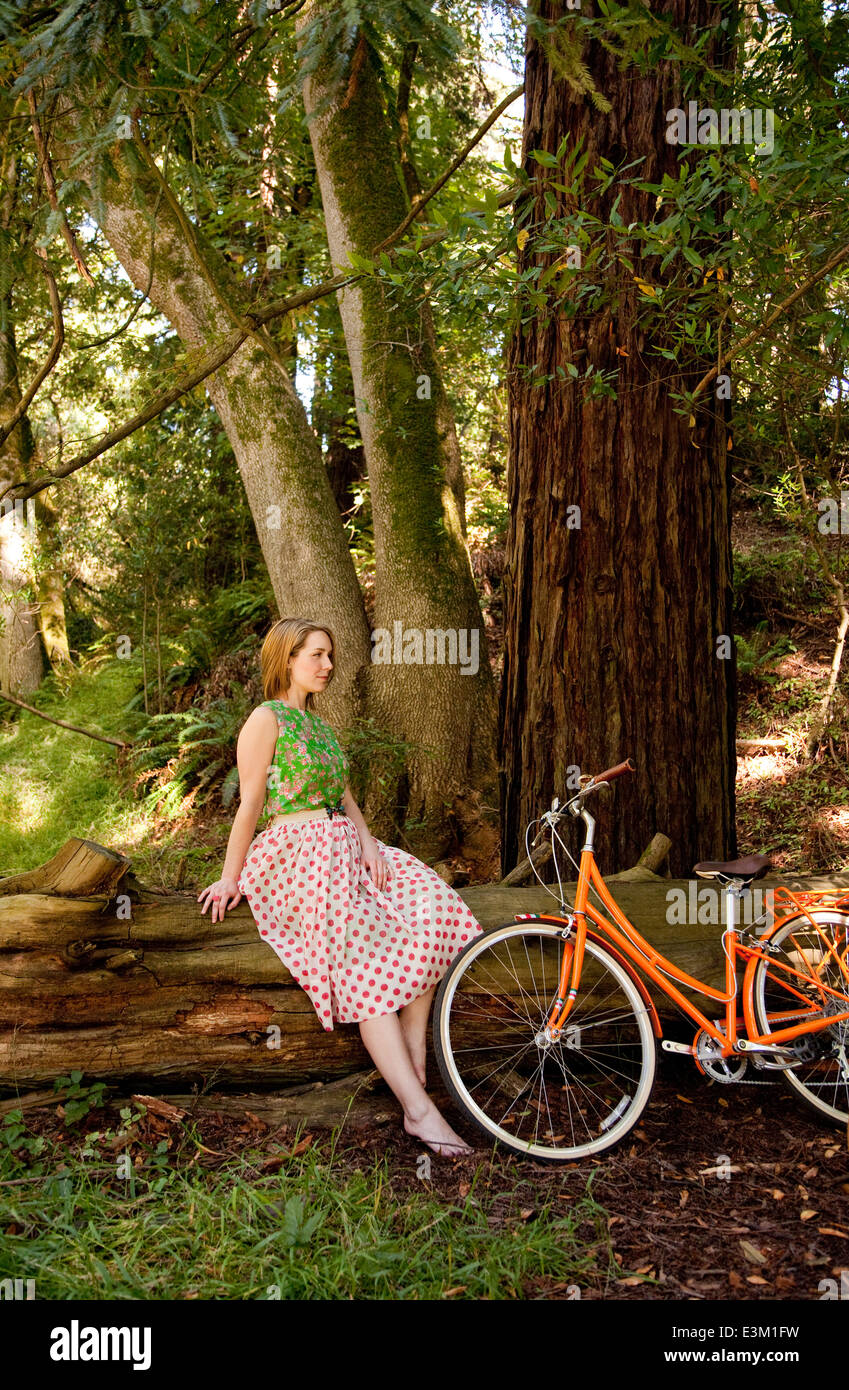 Donna seduta su log in foresta, la bicicletta appoggiata al suo fianco Foto Stock