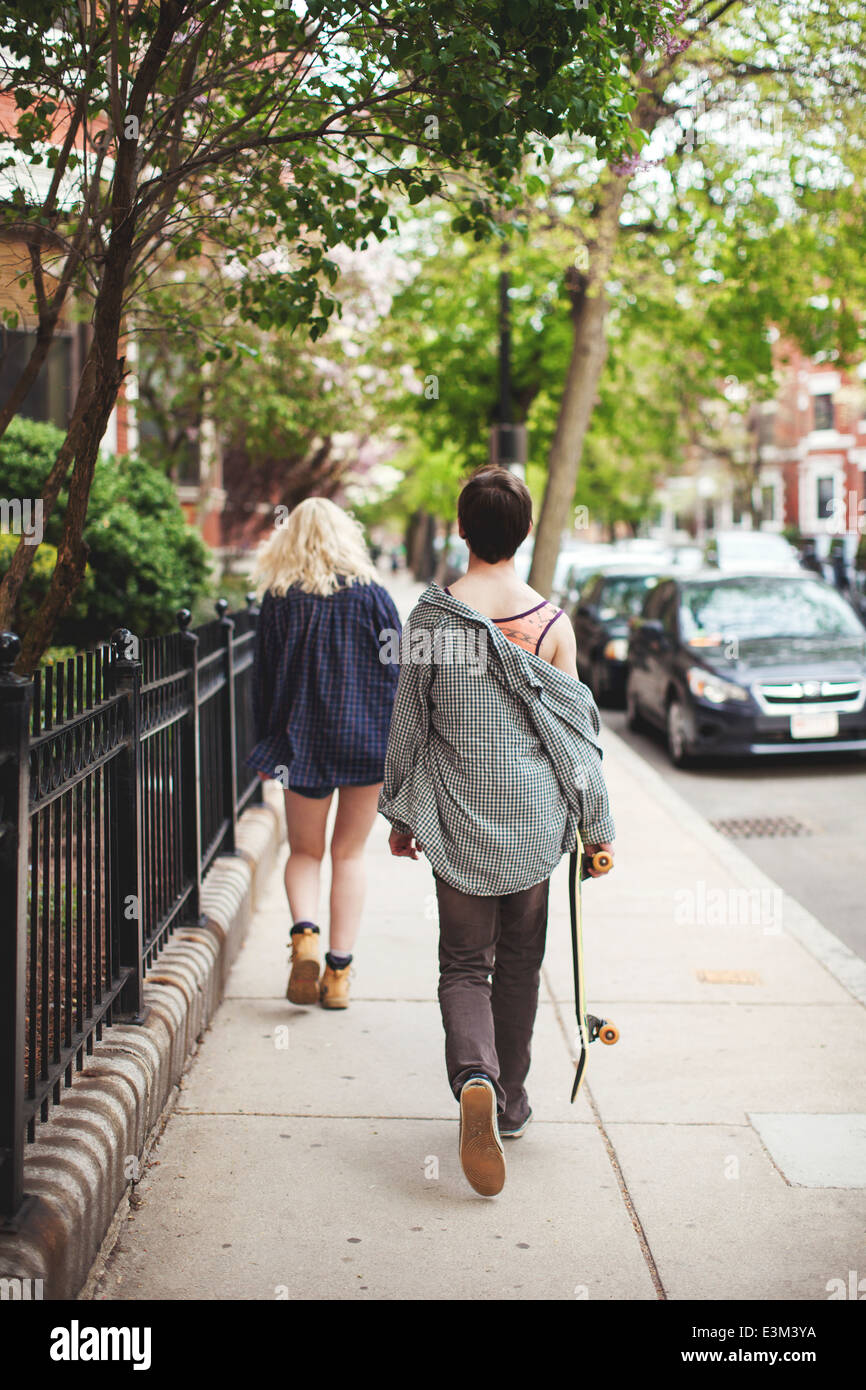 Giovane uomo adulto che trasportano skateboard seguenti giovane donna camminando lungo la strada Foto Stock