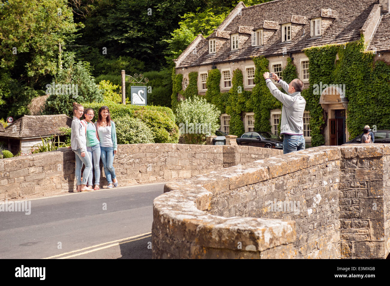 I turisti in posa sul ponte sopra il Fiume Coln & The Swan Hotel nel grazioso villaggio Costwold di Bibury, Gloucestershire, Regno Unito Foto Stock