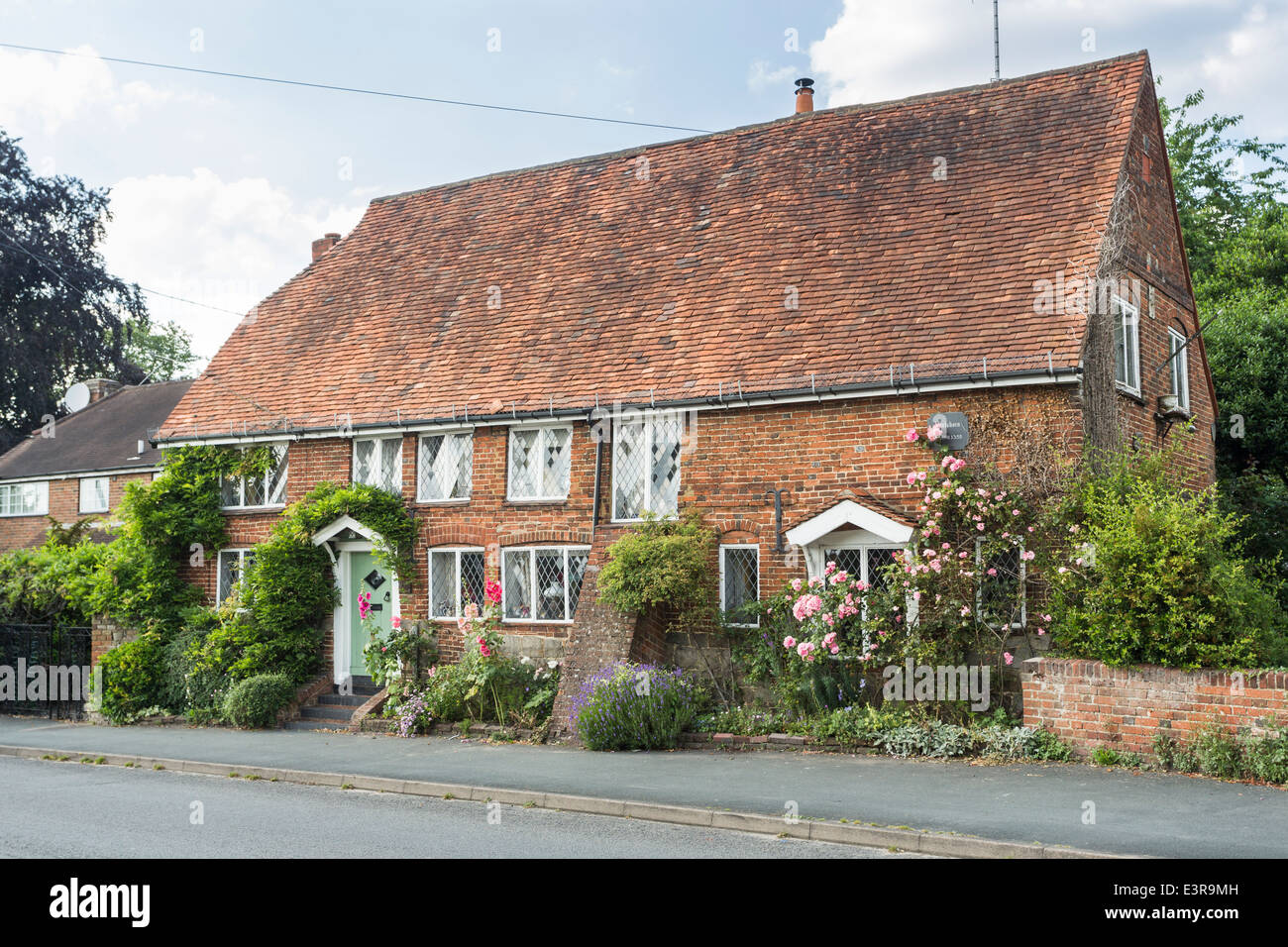Il XIV secolo cottage nel villaggio di cenere vicino a Guildford, Surrey, Regno Unito con hollyhocks e rose attorno alle finestre Foto Stock