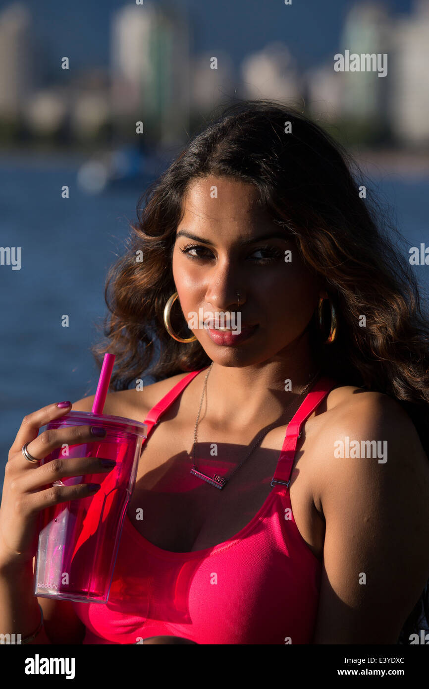 Giovane donna indiana di discesa è di bere da un bicchiere di colore viola e sorridente seductively con l'oceano e gli edifici dietro di lei. Foto Stock