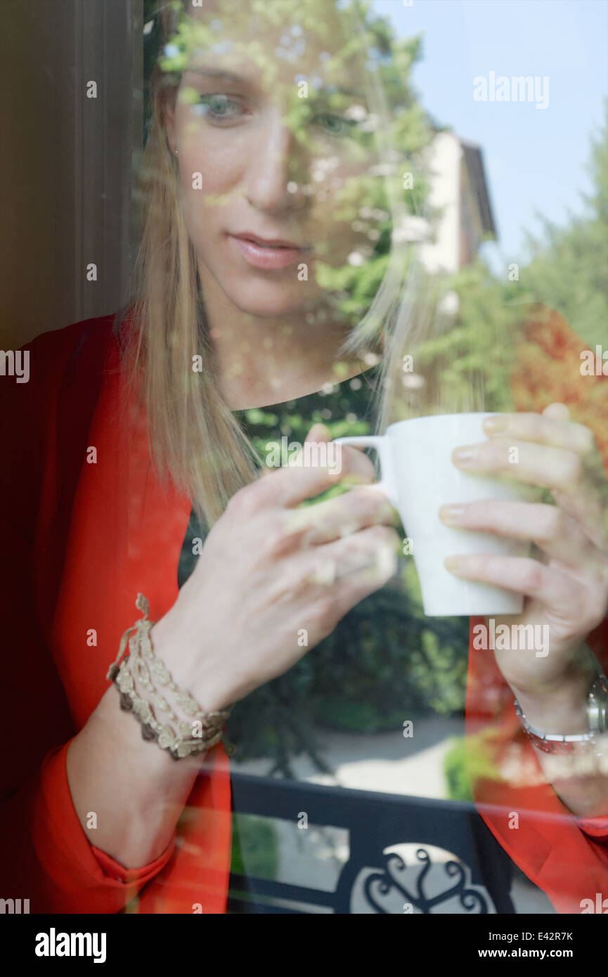 Giovane donna con tazza di caffè guardando fuori della finestra Foto Stock